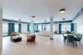 Located at 19 Hemans St in Worcester, Massachusetts, this thoughtfully designed social lounge offers a generous, open-concept layout across a single floor. The main gathering area is anchored by light-toned herringbone hardwood flooring that runs throughout, creating a warm and cohesive aesthetic. Bold navy accent beams contrast with crisp white ceilings, while teal drapery adds rich pops of color across multiple windows that invite abundant natural light and outdoor views.

The lounge is divided into distinct yet seamlessly connected zones. A comfortable seating area features plush cream sofas and mid-century modern leather armchairs arranged around a sculptural coffee table, alongside a striking fireplace wall adorned with hexagonal tile accents. Further into the space, a formal dining area with a large table and upholstered chairs provides an ideal setting for group gatherings. A pool table rounds out the entertainment options, positioned near a windowed corner that overlooks the exterior.

The kitchen is clean and functional, finished with white shaker cabinetry, matte black hardware, white quartz countertops, and a herringbone tile backsplash. It is equipped with a stainless steel refrigerator, sink, and coffee station, making it well-suited to support social events. Flush-mount pendant lighting completes the polished, modern aesthetic throughout this inviting communal space.