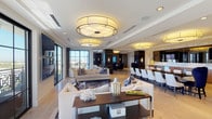 This single-floor luxury space is defined by its open, flowing layout and refined contemporary design. The main living area is anchored by statement drum pendant light fixtures and features light hardwood flooring that extends throughout. Generous seating arrangements, accented with bold blue and ivory tones, create an inviting yet elegant atmosphere, while floor-to-ceiling windows frame panoramic city views and flood the space with natural light.

Adjacent to the living area, a formal dining zone accommodates a large gathering table surrounded by sleek white chairs, seamlessly connected to a fully appointed wet bar. The bar features marble-tiled walls, quartz countertops, stainless steel under-counter refrigeration, and a wall-mounted display screen — ideal for entertaining.

A dedicated media lounge offers a more intimate setting, outfitted with plush navy and cream seating oriented toward a large wall-mounted television, with additional city views visible through a wide-pane window. Dark wood paneling with brass inlay detailing appears as a recurring architectural element, lending warmth and sophistication to the space.

The kitchen area showcases clean-lined wood cabinetry, stone tile backsplash, and integrated appliances, maintaining the space's cohesive modern aesthetic. Throughout, layered lighting — from recessed ceiling fixtures to sculptural pendants and wall sconces — ensures both functionality and ambiance at every turn.