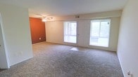 This single-story space centers around an open living area designed to maximize both comfort and functionality. The room is finished with wall-to-wall carpeting in a neutral tone, providing a soft, cohesive foundation throughout. A standout terracotta-colored accent wall adds a warm, bold contrast to the otherwise light and neutral color palette of the space.

Natural light is a defining feature of this living area, with multiple large windows flanking a central exterior door that leads directly to an outdoor area, offering views of the surrounding greenery. The abundance of glazing ensures the interior remains bright and airy throughout the day.

A ceiling fan with an integrated light fixture is centrally positioned overhead, providing both air circulation and ambient lighting. Overhead, a ceiling-mounted HVAC vent suggests a climate control system is in place for year-round comfort. The walls are finished in a clean, light tone that complements the warm accent wall, and baseboard trim runs consistently throughout, adding a neat, finished look to the overall space.