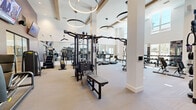 Located at 59 S L St in Livermore, CA, this professionally designed fitness center offers a spacious, single-floor layout that accommodates a full suite of exercise equipment and amenities. The facility is characterized by soaring ceilings that lend an open, airy atmosphere, complemented by large windows that draw in generous natural light and provide views to the outdoors. Striking circular pendant light fixtures add a contemporary aesthetic to the bright, neutral interior.

The main workout floor features light-toned carpet and hardwood flooring in designated zones, keeping the space clean and modern. Floor-to-ceiling mirrors along the walls expand the sense of space and allow users to monitor their form. The cardio section is equipped with a row of treadmills and elliptical machines, each paired with mounted flat-screen televisions for entertainment during workouts. The strength training area includes a multi-station cable machine, adjustable benches, a power rack with free weights, and a selection of plate-loaded and selectorized machines.

A standout feature is a colorful climbing wall, adding a unique and engaging element to the fitness experience. A separate glass-enclosed room provides additional training space, housing stationary bikes, strength machines, and a full dumbbell rack. Structural columns are integrated seamlessly into the layout, contributing to the architectural character of the space without interrupting functionality.