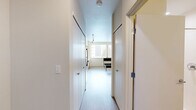 This studio in Seattle, WA is thoughtfully designed to maximize functionality within a compact footprint. The layout is organized around a single open-plan kitchen and living area, where warm wood-toned cabinetry contrasts with crisp white countertops and walls. The kitchen is fully equipped with stainless steel appliances — including a refrigerator, range, built-in microwave, and dishwasher — all illuminated by sleek track lighting overhead. Light wood-tone flooring runs throughout, lending warmth and visual continuity to the space. A large window in the kitchen and living area invites natural light and frames views of the surrounding Seattle cityscape. The entry corridor is flanked by two closets, offering generous storage for a studio of this scale. The bathroom features a warm wood vanity with a white countertop, a large mirror, and a combined shower and tub, maintaining the same consistent design palette found throughout the unit. Clean white walls and neutral finishes give the space an open, airy ambiance that feels both modern and welcoming, making it a highly efficient and practical urban living option.