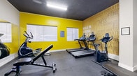Located at 1655 Haro Street in Vancouver's West End, this fitness facility offers a well-equipped exercise room designed to support a range of workout routines. The space makes a strong visual impact with its bold yellow accent walls contrasted against a dark charcoal ceiling and grey rubber flooring, creating an energetic and motivating atmosphere. A large typographic mural featuring fitness-themed words such as "Wellness," "Cardio," "Transform," and "Walk In Strong" covers one wall, reinforcing the space's purpose and adding a dynamic design element.

The exercise room is outfitted with multiple treadmills, stationary bikes, an elliptical trainer, and a cable machine, providing a solid selection of cardio and strength equipment. A dedicated free weights area includes a fully stocked dumbbell rack accompanied by a weight bench, with a wall-length mirror enhancing the sense of space and supporting proper form during workouts. Natural light filters in through several windows along the yellow accent wall, complementing the overhead fluorescent lighting.

The layout is efficient and functional, with clean white walls and white baseboards in the entry area providing a neutral transition into the more vibrantly styled main exercise zone. Safety features including a fire extinguisher are visibly mounted. The facility is accessed through a secure grey panelled door, ensuring controlled entry for authorized users.