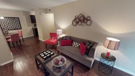 This single-floor residence offers a thoughtfully designed layout that balances comfort and style. The living area serves as the heart of the space, showcasing rich, dark hardwood flooring that adds warmth and a polished finish throughout. A neutral warm-toned color palette on the walls provides a versatile backdrop, complemented by bold red and black accent pieces that create a striking visual contrast.

The living room is furnished with a plush gray sofa adorned with red and patterned throw pillows, alongside a vibrant red accent chair that adds a pop of color. A sleek glass-top coffee table with a dark frame anchors the seating area, adding a modern touch. Decorative wall art featuring interlocking circles enhances the ambiance with an artistic focal point.

The space flows openly toward the kitchen area, which is visible in the background and features dark cabinetry, hinting at a cohesive design language carried throughout the residence. Recessed transitional spaces and defined entryways contribute to a structured yet open feel.

Overall, the space presents a contemporary aesthetic with careful attention to décor details, making it a comfortable and visually engaging single-floor home.
