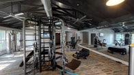 This single-story amenity facility combines functionality with refined design across several distinct areas. The main lounge features soaring ceilings, elegant chandelier lighting, and large dark-framed French doors that flood the space with natural light. Cream-toned sofas, accent chairs, and a dining table create a welcoming, versatile gathering environment with a coastal-inspired color palette of soft blues and neutrals.

A cozy seating nook anchored by a floor-to-ceiling stacked stone fireplace with a rustic wood mantle provides a warm, relaxed atmosphere. French doors adjacent to the fireplace open to an outdoor area, enhancing the sense of connectivity between interior and exterior spaces.

The fitness center is outfitted with professional-grade TKO multi-station strength equipment, cable machines, benches, cycling equipment, and functional training tools including a tire. The room features vaulted ceilings with exposed dark beams, industrial-style pendant lighting, large ceiling fans, mirrored walls, and durable wood-look flooring. Exposed ductwork adds an industrial character that complements the dark trim detailing found throughout the facility, tying together the overall design aesthetic.