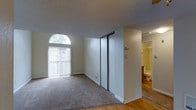 This single-floor residence offers a straightforward and functional layout designed for comfortable everyday living. The main living area is finished with wall-to-wall carpet, providing a warm and soft underfoot feel, while hardwood-style flooring transitions into adjacent spaces, adding visual contrast and continuity throughout the home. A prominent arched window serves as a standout architectural detail, flooding the living space with natural light and lending a classic, elegant touch to the otherwise clean and neutral interior. Vertical blinds offer privacy control while still allowing light to filter through. A set of sliding closet doors provides generous built-in storage, keeping the space organized and uncluttered. The textured ceiling adds subtle depth and dimension to the rooms. Adjacent to the living area, a full bathroom is visible, featuring a toilet, vanity, and shower or tub combination, with warm-toned finishes and tile work that complement the overall neutral palette of the home. The space as a whole maintains a bright, airy ambiance with light-colored walls throughout, creating a clean and inviting atmosphere well-suited for a variety of living arrangements.