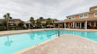 Located at 501 Burke Dr in Hinesville, Georgia, this community amenity space serves as a central gathering hub for residents. The focal point is a generously sized outdoor swimming pool with clear blue water, surrounded by an expansive paver-stone deck that provides ample room for lounging and recreation. A pool volleyball net is set up, adding a recreational element to the aquatic area. Poolside lounge chairs are thoughtfully arranged along the deck, offering comfortable spots for relaxation.

The two-story clubhouse anchors the space with a warm, Mediterranean-inspired architectural style, featuring a terracotta and earth-tone exterior, metal roofing, and arched transom windows. A covered rear porch runs along the building's pool-facing facade, furnished with seating and shaded tables — ideal for outdoor dining or socializing. The clubhouse's upper level features additional windows that allow abundant natural light to fill the interior. 

Mature palm trees and manicured landscaping are interspersed throughout the property, lending a resort-like atmosphere. A shaded pergola structure with additional seating is visible to one side, complementing the overall outdoor living experience. The entire area is enclosed by ornamental fencing, ensuring a secure and private environment for residents to enjoy.