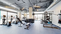Located at 2590 31st Street in Denver, Colorado, this fitness facility offers a generously proportioned, single-floor space designed to accommodate a comprehensive range of workout needs. The interior design follows an industrial-modern aesthetic, characterized by an exposed ceiling with visible ductwork, structural columns clad in light wood paneling, and linear pendant lighting that illuminates the floor below.

The main workout area features durable blue rubber flooring throughout, providing a practical and resilient surface suited to high-intensity activity. Large floor-to-ceiling windows line multiple walls, flooding the space with natural light and offering open views of the surrounding Denver streetscape. This abundance of glazing creates a bright, airy atmosphere that enhances the overall workout environment.

Equipment is organized thoughtfully across the floor. Cardio machines, including treadmills, ellipticals, and stair climbers, are positioned near the windows to take advantage of natural light and outdoor views. A dedicated free weights section features a full rack of dumbbells alongside adjustable benches. A strength training zone includes a multi-function power rack stocked with kettlebells, weight plates, and barbells. A wall-mounted cable machine adds further versatility.

The entry area includes glass double doors, a water station, and built-in wooden shelving, providing a clean and functional transition into the training floor.