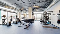 Located at 2590 31st Street in Denver, Colorado, this fitness facility offers a generously proportioned, single-floor space designed to accommodate a comprehensive range of workout needs. The interior design follows an industrial-modern aesthetic, characterized by an exposed ceiling with visible ductwork, structural columns clad in light wood paneling, and linear pendant lighting that illuminates the floor below.

The main workout area features durable blue rubber flooring throughout, providing a practical and resilient surface suited to high-intensity activity. Large floor-to-ceiling windows line multiple walls, flooding the space with natural light and offering open views of the surrounding Denver streetscape. This abundance of glazing creates a bright, airy atmosphere that enhances the overall workout environment.

Equipment is organized thoughtfully across the floor. Cardio machines, including treadmills, ellipticals, and stair climbers, are positioned near the windows to take advantage of natural light and outdoor views. A dedicated free weights section features a full rack of dumbbells alongside adjustable benches. A strength training zone includes a multi-function power rack stocked with kettlebells, weight plates, and barbells. A wall-mounted cable machine adds further versatility.

The entry area includes glass double doors, a water station, and built-in wooden shelving, providing a clean and functional transition into the training floor.