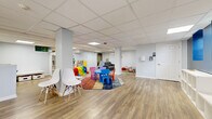 This single-level space is thoughtfully designed as a children's activity and play center, offering a clean, well-organized environment suited for interactive programming and supervised play. The open main area features durable luxury vinyl plank flooring throughout, drop-panel ceilings with recessed lighting, and light gray walls that create a bright, neutral backdrop.

The central play zone is anchored by a colorful child-sized table and chair set atop a multi-colored area rug, surrounded by age-appropriate toys including a crawl tunnel and activity structures. A seating area with a gray sofa and cushioned elements provides a comfortable retreat, while a small private room—furnished with a round white table and modern white chairs—offers a quieter setting for small-group interactions or consultations.

Additional features include a giant Connect Four game station, open shelving units for storage, and wall-mounted hand sanitizer dispensers. A bold navy blue accent wall near the exterior exit adds a pop of color and houses a wall-mounted mini-split HVAC unit. The exterior door provides views of a neighboring green space, allowing natural light to filter into the entry area.