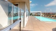 This rooftop amenity space delivers a sophisticated, resort-inspired experience elevated above a vibrant urban skyline. The centerpiece is a generously sized lap pool, framed by wide-plank wood-look tile decking that extends across the entire outdoor level, creating a seamless and polished aesthetic throughout.

Lining the sun deck is a row of lounge chairs positioned to capture maximum sunlight and sweeping panoramic city views, with recognizable landmarks and hillside terrain visible on the horizon. Circular, cushioned sectional seating arrangements with accent pillows are thoughtfully placed along the deck, offering intimate gathering spots that balance comfort with a contemporary design sensibility.

Floor-to-ceiling glass walls and frameless glass barriers run along the building's perimeter, ensuring unobstructed sightlines both from inside and out. Access points feature sleek, stainless steel-handled glass doors with keycard entry, reinforcing the space's secure yet open atmosphere.

A wood-tiled outdoor corridor provides additional circulation around the pool level, while interior-facing glass walls hint at an adjacent fitness or amenity room. The overall ambiance is clean, modern, and resort-caliber, blending open-air relaxation with refined architectural detailing that takes full advantage of its elevated position above the city.