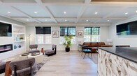 Located at 26 S York Rd in Hatboro, PA, this thoughtfully designed single-floor space offers a refined and welcoming atmosphere ideal for a professional or hospitality setting. The main living area is anchored by a striking built-in wall unit featuring open shelving, a wall-mounted television, and a sleek linear electric fireplace, all framed by soft sage-toned walls that lend a calm, contemporary feel. Elegant coffered ceilings with recessed lighting add architectural depth and a sense of elevated craftsmanship throughout. Light-toned hardwood flooring runs seamlessly across the space, enhancing its bright and airy character. A seating area near large windows allows abundant natural light to fill the room, complemented by tasteful furnishings including modern lounge chairs and a leather sofa. The standout feature is a dramatic wet bar area adorned with a bold blue marble-faced island, a richly veined stone accent wall, wood-paneled tray ceiling, and a sculptural gold chandelier — creating a luxurious focal point. The space balances comfort and sophistication, making it equally suited for client-facing interactions or relaxed gatherings.