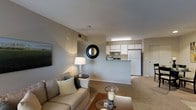 This single-floor residential unit offers a well-planned open-concept layout that connects the living area and kitchen in a comfortable, functional design. The living room features neutral-toned carpeted flooring, smooth walls in a soft gray palette, and a ceiling with recessed ventilation. The space is furnished with a plush beige sofa accented with decorative pillows, a glass-top coffee table, and a side table with a classic table lamp, creating a warm and inviting atmosphere. A decorative round black-framed mirror adds a stylish focal point to the main wall, complemented by framed artwork. The kitchen is accessible via a raised breakfast bar counter that provides a natural visual divider without closing off the space. White cabinetry lines the kitchen walls, offering ample storage, while a full-size white refrigerator and under-cabinet lighting contribute to a clean, practical culinary space. The transition between rooms is smooth and open, enhancing the overall sense of space. Additional doors visible from the main area suggest access to private rooms or storage, rounding out this well-organized and comfortable single-level home.