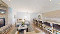 Located at 1090 Summersweet Dr in New Bern, NC, this single-story community clubhouse offers a thoughtfully designed open-plan interior finished in a clean, contemporary coastal palette. Light wood-look flooring runs throughout, complementing soft blue-gray walls and recessed lighting overhead. The welcoming entry foyer features a rustic wood console table and arched mirror, setting a warm tone upon arrival.

The main gathering area centers on a striking floor-to-ceiling stacked stone fireplace with a mounted television above, surrounded by comfortable lounge seating arranged over a patterned area rug. This space flows seamlessly into a well-appointed kitchen and bar area, where an expansive granite-topped island with seating faces a full run of textured flat-front cabinetry, a subway tile backsplash, and an undermount sink. An extended granite counter along the adjacent wall provides generous service space with additional cabinetry above and below.

A dedicated dining nook, filled with natural light from large windows, offers a more intimate setting with a round marble-top table. Large glass doors open directly to an outdoor pool and lounge area, creating a strong indoor-outdoor connection. Ductless mini-split HVAC units ensure year-round comfort throughout the space.