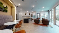 This thoughtfully designed single-floor amenity space combines a fully equipped kitchen, casual dining area, and comfortable lounge into one seamless open-plan layout. Large-format tile flooring in a warm neutral tone runs throughout, unifying each zone and contributing to the spacious, airy feel of the interior.

The kitchen is a standout feature, anchored by a dramatic dark marble island with bar seating and complemented by sleek white cabinetry accented with brushed gold hardware. A textured tile backsplash adds visual depth, while a wood-topped prep counter and stainless steel appliances complete the well-appointed space. Striking geometric gold pendant lights hang overhead, adding a bold design statement.

Adjacent to the kitchen, a casual dining nook features a round white table surrounded by warm cognac-toned chairs with gold-tipped legs, creating an inviting spot for small gatherings. An illuminated wood-paneled accent wall provides a warm, contemporary backdrop.

The lounge area is furnished with rounded, earth-toned swivel chairs arranged on a rich rust-colored area rug, encouraging conversation and relaxation. Floor-to-ceiling glass doors and windows flood the space with natural light and open directly to an outdoor courtyard, offering views of a landscaped pool and patio area. A colorful abstract artwork punctuates the wall, adding a creative finishing touch to this well-curated communal environment.
