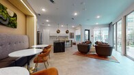 This thoughtfully designed single-floor amenity space combines a fully equipped kitchen, casual dining area, and comfortable lounge into one seamless open-plan layout. Large-format tile flooring in a warm neutral tone runs throughout, unifying each zone and contributing to the spacious, airy feel of the interior.

The kitchen is a standout feature, anchored by a dramatic dark marble island with bar seating and complemented by sleek white cabinetry accented with brushed gold hardware. A textured tile backsplash adds visual depth, while a wood-topped prep counter and stainless steel appliances complete the well-appointed space. Striking geometric gold pendant lights hang overhead, adding a bold design statement.

Adjacent to the kitchen, a casual dining nook features a round white table surrounded by warm cognac-toned chairs with gold-tipped legs, creating an inviting spot for small gatherings. An illuminated wood-paneled accent wall provides a warm, contemporary backdrop.

The lounge area is furnished with rounded, earth-toned swivel chairs arranged on a rich rust-colored area rug, encouraging conversation and relaxation. Floor-to-ceiling glass doors and windows flood the space with natural light and open directly to an outdoor courtyard, offering views of a landscaped pool and patio area. A colorful abstract artwork punctuates the wall, adding a creative finishing touch to this well-curated communal environment.