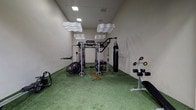 This single-floor fitness room is thoughtfully designed to support a comprehensive workout experience within a compact, focused environment. The space is finished with vibrant green artificial turf flooring, which provides a functional surface suitable for dynamic training, stretching, and functional movement exercises.

At the center of the room stands a multi-functional cable and resistance training rig, equipped for a wide range of strength and conditioning exercises. A heavy-duty punching bag is suspended nearby, adding a combat fitness element to the training options available. Additional equipment includes a fan bike for high-intensity cardio, a dumbbell rack with weights, and an adjustable workout bench, ensuring a well-rounded selection of training tools.

Overhead, the room features recessed ceiling lighting alongside distinctive wave-shaped acoustic panels suspended from the ceiling. These panels contribute both to the room's modern aesthetic and to sound management within the space. The neutral-toned walls keep the environment clean and uncluttered, allowing the equipment to remain the focal point. Overall, the space presents a purpose-built, efficiently organized private gym.
