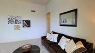 This single-floor space offers a welcoming and well-designed interior that balances comfort with modern style. The living area immediately stands out with its impressively high ceilings that amplify the sense of openness, while the crisp white walls and light neutral carpet create a bright, airy atmosphere throughout. A dark espresso-toned sofa anchors the seating area, complemented by decorative throw pillows featuring a contemporary black, white, and yellow pattern that adds a subtle pop of color. A large black-framed mirror adorns one wall, enhancing the sense of depth and light in the room, while tasteful wall art contributes to the space's refined character. A woven coffee table with decorative glass orbs and a wooden bowl adds texture and warmth to the living area. Overhead, a ceiling vent indicates a central HVAC system for year-round climate comfort. A doorway visible from the living area suggests additional rooms or closet space beyond, hinting at a practical and functional layout. Overall, the space presents a clean, cohesive design with thoughtful details, making it an inviting and comfortable environment.