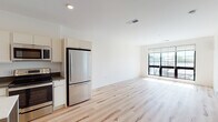 This thoughtfully designed single-floor unit offers a clean, modern aesthetic throughout. The open-concept living and kitchen area is anchored by light-toned hardwood flooring that runs seamlessly across the space, creating a bright and airy atmosphere. Large floor-to-ceiling windows flood the living area with abundant natural light and provide views of the surrounding neighborhood, enhancing the sense of openness.

The kitchen is well-appointed with white flat-front cabinetry, dark countertops, and a full suite of stainless steel appliances, including a refrigerator, electric range, and built-in microwave. Recessed lighting above complements the natural light, making the kitchen a functional and inviting space.

The bedroom is a quiet retreat, finished with plush neutral carpeting and its own large window that continues the theme of natural light throughout the unit. A closet provides convenient built-in storage.

The bathroom maintains the unit's minimalist design language, featuring a floating white vanity with integrated storage drawers and a tall cabinet tower, a wide mirror with overhead lighting, and tile flooring. Clean lines and neutral tones give the space a fresh, contemporary feel.

Overall, the unit presents a cohesive modern design with quality finishes, practical layouts, and an emphasis on light and openness.