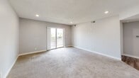 This single-floor, one-bedroom, one-bathroom unit offers an efficiently organized layout across a compact but functional footprint. The living room serves as the central gathering space, finished with plush carpet, recessed lighting, and a textured ceiling. A sliding glass door floods the room with natural light and opens onto a private balcony, providing outdoor views. An open passage connects the living area directly to the kitchen, which features dark espresso cabinetry, light countertops, a stainless steel refrigerator, range, and built-in microwave, all set above durable hardwood-style flooring. A hallway leads from the kitchen toward the bedroom, bathroom, and closet, maintaining a clear circulation path throughout the unit. The bedroom is a well-proportioned, carpeted room with a window allowing in natural light and ample wall space for furnishings. The bathroom includes a full tub-shower combination with built-in shelving, set alongside a vanity with dark cabinetry consistent with the kitchen's design. A dedicated closet provides additional storage. Neutral gray walls and white trim throughout contribute to a clean, cohesive aesthetic.