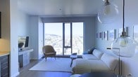 This single-floor residence offers a sleek and contemporary living experience defined by clean lines, modern finishes, and an abundance of natural light. The living area serves as the centerpiece of the space, anchored by expansive floor-to-ceiling windows that frame breathtaking panoramic views of the cityscape and rolling hills beyond — a truly standout architectural feature that floods the room with daylight and creates a seamless connection between the interior and the urban landscape outside.

The living room is thoughtfully appointed with a plush white sectional sofa, a curved accent chair in warm tan leather, and a minimalist coffee table arranged atop a textured area rug, creating an inviting yet refined atmosphere. A sculptural pendant light fixture adds an artistic focal point overhead, complementing the room's modern aesthetic. The walls feature carefully curated artwork, adding subtle color and personality to the otherwise neutral palette of whites, grays, and warm wood tones found in the flooring.

The overall ambiance is sophisticated and airy, with the high-rise vantage point and generous glazing ensuring that natural light and city views remain constant companions throughout the space. The design balances openness and comfort, making it both visually striking and functionally inviting.