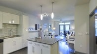 This single-floor residence showcases a thoughtfully designed open-concept layout that seamlessly connects the kitchen, dining, and living areas into one cohesive and inviting space. The modern kitchen is anchored by a central island with a sleek dark stone countertop and crisp white cabinetry with brushed chrome hardware. Elegant pendant lights hang above the island, complemented by a track lighting fixture that highlights the workspace below. The kitchen flows naturally into a dedicated dining area, where a glass-top table is surrounded by white upholstered chairs, creating a refined yet approachable setting. Beyond the dining area, the living room offers a comfortable and well-appointed retreat, decorated in a coordinated blue and white palette with patterned rugs and accent pieces. Rich dark hardwood flooring runs throughout, grounding the bright, airy aesthetic. A ceiling fan with a light fixture adds both function and style to the living area. Large windows dressed with white floor-to-ceiling curtains allow abundant natural light to fill the space, while providing views of the surrounding greenery. The overall ambiance is clean, contemporary, and cohesive, making this space both visually striking and highly functional.