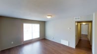 This single-floor space presents a straightforward and functional layout designed for comfortable everyday living. The main living area features rich, warm-toned hardwood flooring that extends throughout, adding a sense of warmth and continuity to the space. Neutral gray-beige walls provide a versatile backdrop that complements a variety of furnishing styles and décor choices.

A large window fitted with horizontal blinds allows abundant natural light to fill the room, creating a bright and open atmosphere. The textured ceiling adds a subtle dimension to the overhead space, while a centrally mounted flush light fixture provides reliable ambient illumination.

A wall-mounted baseboard heater ensures efficient climate control, and a thermostat panel is conveniently positioned on the wall, offering easy temperature management. Visible ductwork vents suggest a broader HVAC system is in place throughout the unit.

The clean lines, minimal detailing, and practical finishes throughout the space make it well-suited for those seeking a move-in ready environment. The open, uncluttered design allows for flexible furniture arrangement and personalization. Overall, this space offers a calm, neutral setting with key functional features that support comfortable, day-to-day living.