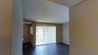 This single-floor space presents a clean, functional layout centered around an open living area. The room is anchored by dark hardwood-style flooring that contrasts with the warm, neutral-toned walls, creating a grounded and cohesive aesthetic throughout. A textured ceiling adds subtle visual depth to the space.

The most prominent feature of the living area is a large sliding glass door dressed with vertical blinds, which serves as the primary source of natural light. The door allows an abundance of daylight to filter into the room, creating a bright and airy atmosphere. An additional window is visible in the adjoining area, further enhancing the natural light throughout.

The open layout is defined by a wide entryway or pass-through that connects adjacent areas of the unit, lending a sense of spaciousness and flow. White baseboards provide a clean finish along the perimeter of the room, complementing the neutral color palette. The overall ambiance of the space is calm and understated, offering a versatile backdrop well-suited for a variety of furnishing styles and arrangements.