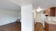 This two-story residence offers a practical and well-organized layout across two floors. The main level features rich, dark hardwood-style flooring that runs throughout the open living areas, creating a warm and cohesive aesthetic. A central wall divides the space, separating the living area from the kitchen corridor, providing a sense of structure and definition within the floor plan.

The kitchen is positioned along one side of the main level and is equipped with white appliances, including a full-size refrigerator and range. Light-toned cabinetry lines the kitchen walls, offering ample storage, while the hardwood-style flooring continues seamlessly into this space. Overhead lighting provides functional illumination throughout.

A carpeted staircase, visible from the main living area, leads to the upper floor, adding a soft textural contrast to the hard flooring below. The staircase is neatly integrated into the layout, with the lower landing partially visible and offering a glimpse into the additional space above.

Throughout the home, clean white walls and ceilings contribute to a bright, neutral atmosphere that feels open and easy to personalize. The overall design is straightforward and functional, making it well-suited for everyday living across both levels.