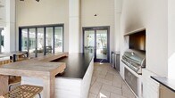 Located at 1931 Chemin Metairie Pkwy in Youngsville, Louisiana, this single-level amenity space delivers a resort-caliber outdoor experience designed for both relaxation and socializing. The centerpiece is a generously sized swimming pool with crystal-clear blue water, featuring in-pool lounge ledges with sun chairs and an ADA-compliant pool lift to ensure accessibility for all residents. The pool deck is lined with sleek chaise lounges and surrounded by thoughtfully arranged outdoor seating areas. Adjacent to the pool, a covered outdoor kitchen and entertainment pavilion stands out with its dramatic soaring ceilings, white shiplap walls, and open-air design that invites natural light and breeze. The kitchen area is outfitted with a built-in stainless steel grill, ample counter space with dark stone surfaces, a white brick base, and bar-height seating with woven rattan chairs — ideal for casual gatherings. Multiple high-top tables are positioned beneath the covered pavilion, offering views of the pool. Outdoor shower stations and manicured landscaping with native palms and subtropical plantings complete the exterior. The overall architectural style blends contemporary Southern design with clean white facades, metal roofing accents, and dark-trimmed windows, creating an inviting and polished community retreat.