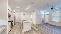 This well-appointed single-floor residence offers a clean, contemporary design highlighted by light wood-style flooring and crisp white walls that carry consistently throughout the space. The open-concept layout places the kitchen at the heart of the home, where a central island with a granite countertop and double sink serves as both a functional workspace and a natural gathering point. The kitchen is fully equipped with stainless steel appliances, including a gas range, over-the-range microwave, and a full-size refrigerator, all complemented by white shaker-style cabinetry with ample storage and open shelving. Recessed lighting illuminates the kitchen and adjoining areas, adding a polished, modern feel. The living area benefits from natural light streaming through large windows dressed with blinds, and a ceiling fan adds comfort to the open space. Bedrooms are generously sized with clean white walls and the same wood-style flooring, offering a bright and versatile canvas. A walk-in closet with wire shelving provides practical storage. The unit also includes a dedicated in-unit laundry area with a full-size washer and dryer. The overall ambiance is modern, light-filled, and move-in ready.