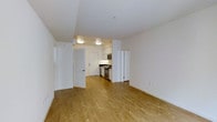 This single-floor unit showcases a bright, open-concept layout that seamlessly connects the living area to the kitchen. The space is finished with warm-toned hardwood flooring that runs throughout, contributing to a cohesive and inviting atmosphere.

The kitchen is set within a defined nook, featuring stainless steel appliances including a gas range, microwave, and refrigerator, paired with white cabinetry and dark countertops. Flush-mounted ceiling lights provide consistent, functional illumination across both the kitchen and main living area.

The living space is generously proportioned, offering flexibility for various furniture arrangements. White walls and a smooth white ceiling enhance the sense of openness and brightness throughout the unit. Two interior doors are visible — one leading to what appears to be a closet or utility space, and another providing access to an additional room, suggesting practical storage and spatial organization.

Overall, the unit presents a clean, modern aesthetic with neutral finishes that create a versatile and well-lit environment suited to a range of interior styles.