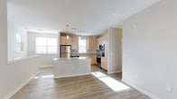 This single-floor, 2-bedroom residence with garage access offers a thoughtfully designed open-concept layout that seamlessly connects the kitchen and living areas. The kitchen is a standout feature, equipped with light maple cabinetry, stainless steel appliances including a refrigerator, range, and microwave, and a generously sized central island topped with speckled granite countertops. Pendant lighting hangs above the island, providing both task illumination and a modern aesthetic touch. Track lighting above the main kitchen workspace adds further functionality.

The open layout flows naturally into the living area, where hardwood-style flooring extends throughout, adding warmth and continuity to the space. A ceiling fan in the living area enhances comfort, while multiple windows along the exterior walls invite abundant natural light, giving the home a bright and airy feel. Neutral gray walls throughout provide a clean, versatile backdrop. The overall ambiance is modern and welcoming, with a design that balances practicality and style across the single level.