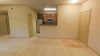 This single-floor space features an open-concept design that connects the kitchen and living area into one cohesive, functional layout. The kitchen is defined by a raised peninsula counter that serves as a natural divider, offering both workspace and a casual seating or serving area. Medium-toned wood cabinetry lines the kitchen walls, providing ample storage, while stainless steel appliances — including a full-size refrigerator and a built-in microwave above the range — add a modern, practical touch. Two pendant lights hang above the peninsula, complementing the flush overhead lighting and contributing to a warm, even illumination throughout the space. The flooring transitions from hard surface tile in the kitchen area to plush carpeting in the living space, adding comfort underfoot. Neutral cream and beige tones on the walls and flooring create a clean, versatile backdrop throughout. A paneled door visible along one wall leads to an additional room, suggesting further private living space beyond the main open area. Overall, the layout feels open and easy to navigate, with a straightforward, functional design suited to everyday living.