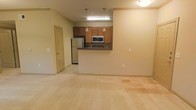 This single-floor space features an open-concept design that connects the kitchen and living area into one cohesive, functional layout. The kitchen is defined by a raised peninsula counter that serves as a natural divider, offering both workspace and a casual seating or serving area. Medium-toned wood cabinetry lines the kitchen walls, providing ample storage, while stainless steel appliances — including a full-size refrigerator and a built-in microwave above the range — add a modern, practical touch. Two pendant lights hang above the peninsula, complementing the flush overhead lighting and contributing to a warm, even illumination throughout the space. The flooring transitions from hard surface tile in the kitchen area to plush carpeting in the living space, adding comfort underfoot. Neutral cream and beige tones on the walls and flooring create a clean, versatile backdrop throughout. A paneled door visible along one wall leads to an additional room, suggesting further private living space beyond the main open area. Overall, the layout feels open and easy to navigate, with a straightforward, functional design suited to everyday living.