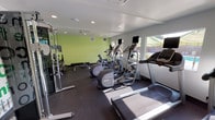 This fitness center is thoughtfully designed to support a well-rounded workout experience. The space is finished with light green accent walls and a bold motivational word mural — featuring phrases like "strength," "commitment," and "inspire" — creating an energizing atmosphere throughout. Recessed lighting and a ceiling-mounted HVAC unit keep the environment comfortable and well-lit.

The cardio section is generously equipped with multiple treadmills, elliptical trainers, and upright and recumbent stationary bikes, each outfitted with individual display monitors. Rubber flooring throughout provides both safety and durability underfoot.

For strength training, the facility includes a multi-functional cable machine, a dedicated free weight area with a full rack of dumbbells in varying sizes, and a flat workout bench. Natural light filters in through windows and a large glass door along one wall, offering views of an outdoor pool and patio area — a pleasant visual connection to the surrounding amenity spaces.

The single-floor layout is open and efficiently organized, allowing easy movement between equipment zones. Overall, the fitness center presents a clean, modern, and motivating environment suited for a variety of fitness routines.