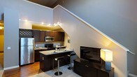 This two-story space is defined by its striking vertical scale, with soaring ceilings that create an airy, open atmosphere throughout the main level. The dramatic staircase is a central architectural feature, rising prominently along one wall and drawing the eye upward while reinforcing the sense of height and volume.

The open-concept main level integrates the kitchen and living areas seamlessly. The kitchen is outfitted with dark espresso cabinetry, granite countertops, and stainless steel appliances, including a refrigerator and microwave. A kitchen island with bar-height seating serves as both a functional workspace and a casual dining area. Track lighting above the kitchen provides focused, warm illumination, enhancing the rich tones of the cabinetry and countertops.

The living area sits adjacent to the kitchen and features hardwood flooring that transitions into carpet, adding texture and warmth. A large flat-screen television and media console anchor this space. The overall ambiance is modern and urban, characterized by a neutral color palette of gray and warm white tones complemented by the deep wood finishes throughout.