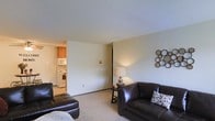This single-floor space offers a straightforward and functional layout, with the living area serving as the central gathering space. The room features wall-to-wall carpeting in a neutral tone, creating a comfortable and cohesive foundation throughout. The living area is furnished with dark leather seating, including a sofa and chaise, accented by decorative pillows and a table lamp that adds a warm glow to the space. A circular mirror wall accent provides a modest decorative touch.

The open sightline from the living area leads directly into the kitchen, which is separated by a partial wall, maintaining a sense of openness while defining each functional zone. The kitchen is equipped with white appliances, including a range and over-the-range microwave, and features warm-toned wooden cabinetry that provides ample storage. The light-colored walls throughout the space contribute to a bright and airy feel. The textured ceiling adds a classic finish consistent with the overall design. The layout prioritizes practicality, making efficient use of the available space while maintaining a clean and livable environment.