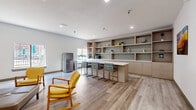 This thoughtfully designed single-floor community space combines functionality with a warm, contemporary aesthetic. The open-plan layout is anchored by light wood-look flooring that runs throughout, creating a cohesive and welcoming atmosphere across all areas.

A standout feature is the built-in shelving wall, finished in warm natural wood tones with a subtle tile backsplash, providing both display space and storage. Adjacent to it, a sleek white-topped counter island serves as a bar-height gathering point, accompanied by modern gold-framed stools with gray upholstered seats. A stainless steel refrigerator and countertop microwave complete the kitchenette area, making it a practical hub for communal use.

The lounge area is furnished with distinctive yellow rocking chairs and complementary seating, arranged to encourage conversation and relaxation. White-topped activity tables with clean metal frames offer flexible workspace throughout the room. Recessed lighting illuminates the space evenly, while multiple windows along the exterior walls invite generous natural light and views of the outdoors.

An open passage connects to an adjacent lounge area featuring plush dark armchairs set on a patterned area rug, with bold yellow ceiling accents adding a vibrant architectural touch. Colorful artwork adorns the walls, contributing to the space's lively and engaging character. The overall environment is bright, accessible, and designed for community interaction.