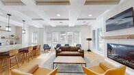 This thoughtfully designed single-floor amenity space blends classic architectural details with contemporary furnishings to create an inviting and functional communal environment. The space is anchored by elegant coffered ceilings that lend a sense of height and sophistication throughout. Underfoot, warm herringbone-patterned hardwood flooring runs seamlessly across the entire area, tying each zone together with a cohesive visual flow.

The main lounge area serves as the centerpiece, featuring a built-in fireplace framed by a rustic wood mantel and shiplap-style paneling, with a wall-mounted television positioned above. Comfortable seating arrangements — including a tufted sofa, accent chairs in warm amber leather, and navy blue lounge chairs — are thoughtfully grouped to encourage conversation and relaxation. Large windows dressed with white blinds allow abundant natural light to fill the space while offering views of the surrounding greenery outdoors.

Adjacent to the lounge, a casual dining area with a round wood table and modern chairs provides an intimate setting for small gatherings. Nearby, a kitchenette bar features a marble-topped island with bar stool seating, pendant lighting, open wood shelving, and a self-serve beverage station with a coffee and drink dispenser. Paneled wainscoting along the walls throughout adds a refined, classic touch to the overall design.