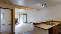 This single-floor space showcases an open-concept design that connects the kitchen and living areas seamlessly. The kitchen features a wrap-around laminate bar counter with a warm wood-tone edge, providing a natural division between the cooking and living spaces. Track lighting overhead adds a functional and modern touch to the kitchen zone. The living area is carpeted and centers around a tile-surround fireplace, lending a cozy focal point to the room. A sliding glass door with vertical blinds allows natural light to filter in and provides access to an outdoor balcony, where views of surrounding greenery are visible. The transition between the kitchen's hard flooring and the living area's carpet is clearly defined, adding visual structure to the open floor plan. Walls are painted in neutral tones throughout, complemented by a textured ceiling finish. The layout is practical and functional, with the open sightlines between spaces creating a sense of spaciousness. Overall, the space offers a comfortable, straightforward design suited for everyday living.