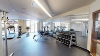This fitness center is a comprehensively equipped facility designed to support a full range of workout routines. The space is housed on a single floor and is characterized by its clean, modern aesthetic, with neutral-toned walls, recessed and linear pendant lighting, and durable speckled rubber flooring throughout that provides both safety and resilience underfoot.

The main workout area is generously proportioned, offering ample room to move between equipment without feeling crowded. Cardio equipment is well-represented, with multiple treadmills, elliptical trainers, and stationary and upright bikes arranged to make efficient use of the floor space. For strength training, the facility includes a multi-function cable and weight rack system, a dedicated free weights section featuring a full dumbbell rack with a wide range of weights, and a weight bench with plates.

Full-length mirrors line one wall, enhancing the sense of openness and allowing users to monitor their form during exercise. Large sliding glass doors and windows along the perimeter flood the space with natural light and provide direct views of the outdoor surroundings, including what appears to be a pool area and exterior courtyard. A separate entry vestibule with warm wood-framed glass doors provides a clean transition into the gym. Overall, the facility offers a bright, functional, and professional environment well-suited for daily fitness use.