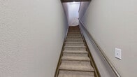 This two-floor residence offers a thoughtfully organized layout across a compact yet functional footprint. The ground level is dedicated primarily to an attached garage with a concrete floor, overhead fluorescent lighting, and an in-unit water heater — providing both parking and additional storage utility. A carpeted staircase with a metal handrail connects the lower level to the main living floor above.

The upper floor hosts the majority of the living space. The open-concept kitchen and living area forms the heart of the unit, where rich, dark hardwood flooring extends throughout. The living area features a ceiling fan and a set of glass-paned French doors that invite natural light and offer outdoor views. The kitchen is well-appointed with rich walnut-toned cabinetry, light granite countertops, a subway tile backsplash, and a full suite of stainless steel appliances.

The bedroom is carpeted and features a ceiling fan, a window with outdoor views, and access to a dedicated walk-in closet with built-in shelving. The bathroom continues the warm aesthetic with hardwood flooring, rich cabinetry, and granite countertops. Six-panel interior doors are used consistently throughout, contributing to a cohesive design. Overall, the space balances practicality with warm, inviting finishes.