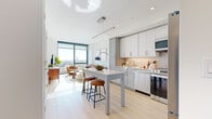 This single-floor residence showcases a thoughtfully designed open-concept layout that seamlessly integrates the kitchen, dining, and living areas. The kitchen is outfitted with two-tone cabinetry—white upper cabinets paired with soft grey lower units—complemented by a neutral subway tile backsplash, stainless steel appliances including a dishwasher, range, microwave, and refrigerator, and a white quartz countertop island with bar seating for two. Light wood flooring runs throughout, reinforcing a clean, cohesive aesthetic.

The living area benefits from expansive floor-to-ceiling windows that frame sweeping city views and flood the space with natural light. The bedroom continues this theme with its own large window offering elevated urban vistas, light wood flooring, and ample room for a desk and seating area. A bathroom accessed via a private corridor features large-format tile flooring, a wall-mounted mirror, and a vanity with white cabinetry and ample storage. Throughout the residence, a neutral white and grey palette, recessed lighting, and modern fixtures contribute to a bright, contemporary ambiance.
