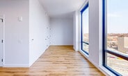 Located at 69-02 Queens Boulevard in Woodside, NY, this thoughtfully designed unit offers a clean, modern aesthetic throughout its single-floor layout. The open-plan kitchen and living area serves as the heart of the space, featuring sleek white flat-front cabinetry extending to the ceiling, light quartz countertops, a matte black faucet, and stainless steel appliances including a professional-style range and a full-size refrigerator. Warm honey-toned hardwood-style flooring runs continuously throughout, complementing the crisp white walls and creating a cohesive, contemporary feel. The living area benefits from a floor-to-ceiling window framed in black, flooding the space with natural light and offering open views of the Woodside skyline. Closet space is provided via a set of clean white double doors with black hardware. The bathroom is a standout feature, finished with large-format marble-look wall tile, a deep soaking tub with an overhead rain showerhead, a built-in niche, a floating wood-toned vanity with a concrete-style countertop, an undermount sink, and matte black fixtures throughout. Recessed lighting and a large mirror complete the refined, spa-like atmosphere of the bathroom.