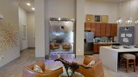This single-floor space makes an immediate impression with its striking floor-to-ceiling indoor water wall, branded with the "Enclave" logo and framed in sleek stainless steel. The cascading water flows over an illuminated base of polished river stones, creating a captivating focal point that blends artistry with ambiance. The surrounding space benefits from high ceilings that amplify the dramatic scale of this installation.

The open-concept layout seamlessly connects the living area to the kitchen. The living space is furnished with curved cognac leather seating and accented by decorative sculptural pieces, lending a contemporary yet inviting feel. Light wood-toned flooring runs throughout, contributing to the warm, cohesive aesthetic.

The kitchen is well-appointed with warm medium-toned wood cabinetry, light countertops, and stainless steel appliances, including a full-size refrigerator. A series of decorative framed panels above the upper cabinets add visual interest. Neutral wall tones and recessed lighting throughout the space ensure a bright, polished atmosphere that balances modern design with comfortable livability.