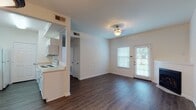 This single-story residence is designed with a practical, open layout that connects the living, dining, and kitchen areas into a cohesive and functional space. Rich wood-look flooring flows throughout the main living areas, complementing the clean white walls and crisp baseboards that define the interior aesthetic.

The living area is anchored by a corner fireplace with a classic white mantel, adding warmth and character to the space. A pendant light fixture and flush-mount ceiling light illuminate the open area, which also benefits from natural light streaming through a windowed exterior door that provides outdoor views. The dining area is served by an additional ceiling light fixture.

The galley-style kitchen is efficiently arranged with white cabinetry, light countertops, and white appliances including a range, over-the-range microwave, refrigerator, and dishwasher. A pass-through opening connects the kitchen visually to the main living area, enhancing the sense of openness.

The bedrooms feature plush carpeting, ceiling fans with lighting, natural light from windows, and closet space. One bedroom includes a walk-in closet and direct access to a full bathroom equipped with a wide vanity, large mirror with vanity lighting, and a shower/tub combination.