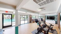 Located at 4301 North 24th Street in Phoenix, AZ, this single-floor fitness center offers a well-appointed and thoughtfully designed space for residents to exercise and unwind. The layout encompasses a main workout area, a lounge, a free weights room, and connecting hallways, all finished with durable cork-style flooring throughout.

The primary exercise area is bright and open, featuring treadmills, Peloton cycling bikes, and interactive fitness screens. Floor-to-ceiling sliding glass doors line one wall, flooding the space with natural light and providing direct access to the outdoor pool area, creating a seamless indoor-outdoor connection.

A dedicated free weights room makes a bold design statement with lush, tropical botanical wallpaper covering every wall, complemented by a mirrored accent and a dumbbell rack — offering an energizing and visually striking environment for strength training.

The lounge area provides a comfortable retreat, featuring a built-in L-shaped wood bench with cushions, a glass-top coffee table, a wall-mounted TV, and large windows that frame views of the surrounding landscaping and pool. The teal color accents found on structural columns and in the hallways tie the entire space together with a cohesive, modern aesthetic. Clean white hallways with tile flooring and contemporary flush lighting connect all areas efficiently.