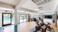 Located at 4301 North 24th Street in Phoenix, AZ, this single-floor fitness center offers a well-appointed and thoughtfully designed space for residents to exercise and unwind. The layout encompasses a main workout area, a lounge, a free weights room, and connecting hallways, all finished with durable cork-style flooring throughout.

The primary exercise area is bright and open, featuring treadmills, Peloton cycling bikes, and interactive fitness screens. Floor-to-ceiling sliding glass doors line one wall, flooding the space with natural light and providing direct access to the outdoor pool area, creating a seamless indoor-outdoor connection.

A dedicated free weights room makes a bold design statement with lush, tropical botanical wallpaper covering every wall, complemented by a mirrored accent and a dumbbell rack — offering an energizing and visually striking environment for strength training.

The lounge area provides a comfortable retreat, featuring a built-in L-shaped wood bench with cushions, a glass-top coffee table, a wall-mounted TV, and large windows that frame views of the surrounding landscaping and pool. The teal color accents found on structural columns and in the hallways tie the entire space together with a cohesive, modern aesthetic. Clean white hallways with tile flooring and contemporary flush lighting connect all areas efficiently.