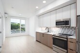 This single-floor unit showcases a clean, contemporary design with light wood-tone flooring and a bright, neutral palette that runs consistently throughout the space. The open-plan layout connects the kitchen directly to the living area, creating a cohesive and functional flow.

The kitchen is a standout feature, fitted with two-tone cabinetry — warm wood lower cabinets paired with matte grey upper cabinets — and accented by matte black hardware. A geometric patterned tile backsplash adds visual interest behind the stainless steel appliances, which include a range with electric cooktop, an over-range microwave, and a full-size refrigerator. White quartz countertops and an undermount sink with a matte black faucet complete the refined look.

The living area receives generous natural light through a large sliding window that frames an urban streetscape, while a wall-mounted mini-split unit provides climate control. The bedroom continues the light, airy aesthetic with its own oversized window offering elevated city views.

The bathroom features white subway tile lining a soaking tub surround, wood-look tile flooring, and coordinating matte black fixtures and accessories, maintaining the modern and cohesive design language found throughout the entire space.