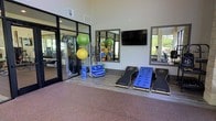 This modern fitness center is designed to accommodate a comprehensive range of workout styles within a clean, open layout. The space is distinguished by soaring ceilings that lend an airy, expansive feel, complemented by large floor-to-ceiling glass doors and windows that invite abundant natural light and provide views of the surrounding greenery. The flooring transitions between durable rubber sport surfacing and wood-toned panels, delineating distinct workout zones effectively. A dedicated functional training area is stocked with Matrix exercise mats, stability balls in multiple sizes, resistance bands, and foam rollers neatly organized on a multi-tier rack. Wall-mounted mirrors amplify the sense of space and allow for form checks during exercise. A flat-screen TV is mounted on the wall, adding an entertainment element to training sessions. Glass-paneled doors open to an adjacent strength training zone, visible in the background and outfitted with additional equipment. The overall ambiance is bright, clean, and welcoming, reflecting a thoughtfully designed environment built to support fitness goals across skill levels.